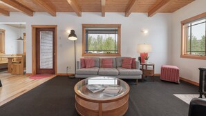 Un salon spacieux avec un plafond en bois, une table basse ronde, un canapé gris agrémenté de coussins à rayures, une petite table d’appoint et une fenêtre donnant sur la forêt.