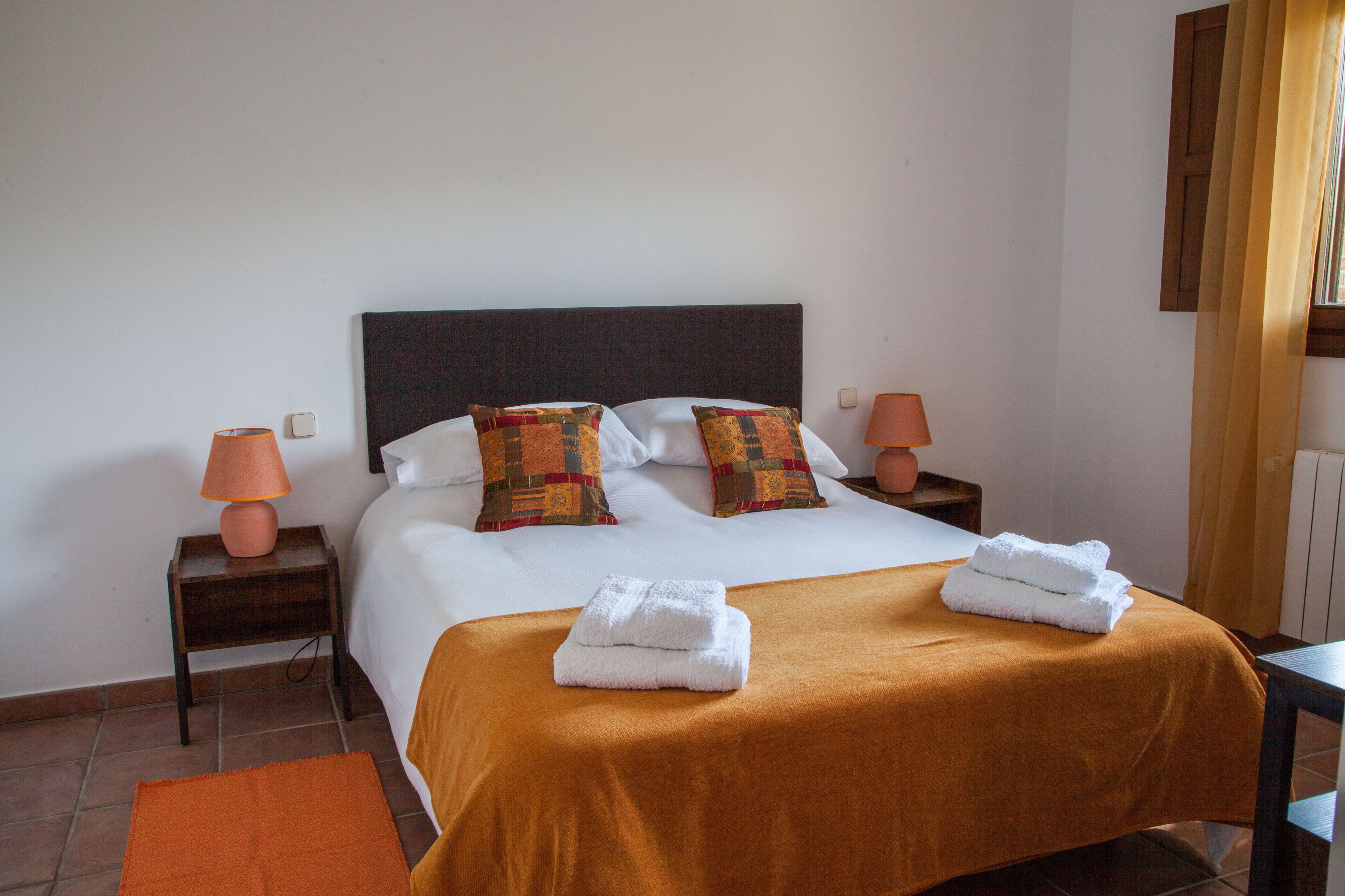 Una cama bien hecha con dos almohadas, una colcha marrón y dos toallas dobladas. Hay dos lámparas de noche y una alfombra naranja pequeña en el suelo.