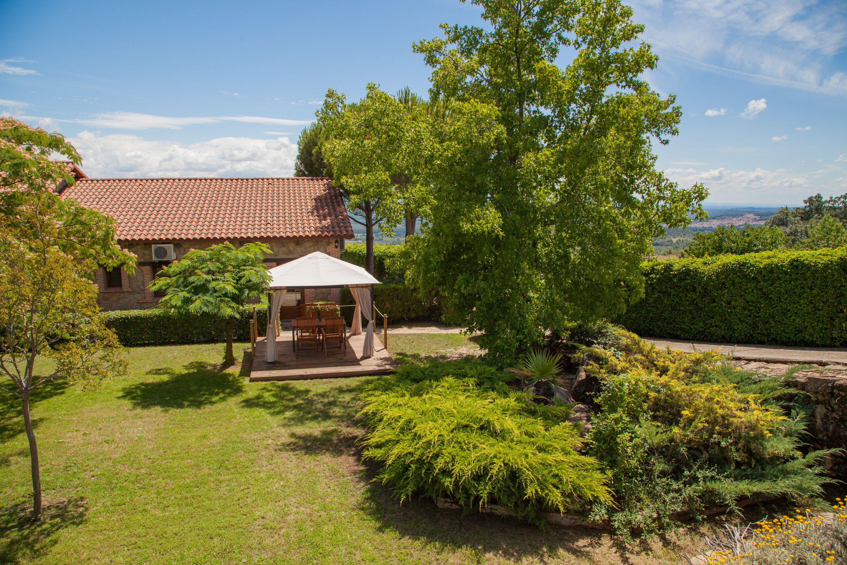 A house with a red-tiled roof, a covered patio with furniture, and a lush garden with various plants and a small pond.