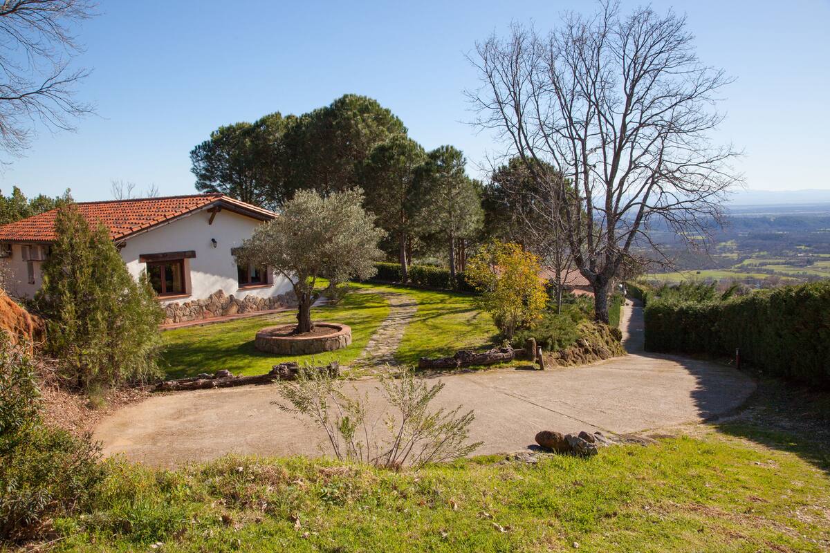A house with a stone foundation, a red-tiled roof, and a well-maintained lawn with a pathway leading to the entrance.