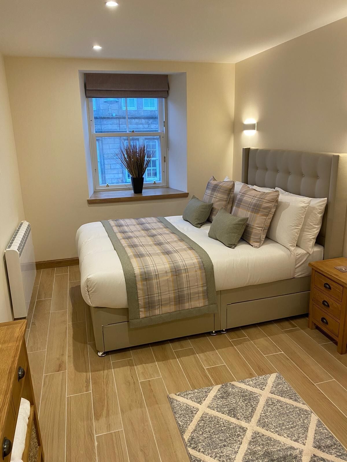 A bedroom with a bed, bedside table, nightstand, radiator, window, and a view of the outside.