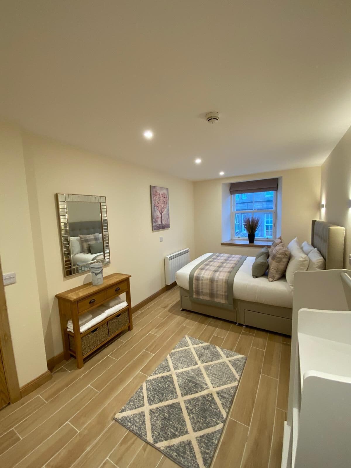 A bedroom with a bed, bedside table, mirror, radiator, window, and a view of the outside.