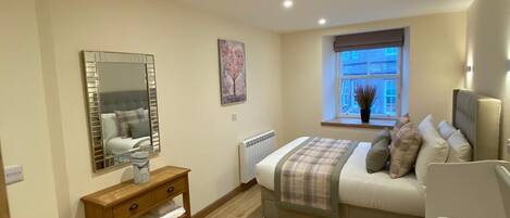 A bedroom with a bed, bedside table, mirror, radiator, window, and a view of the outside.