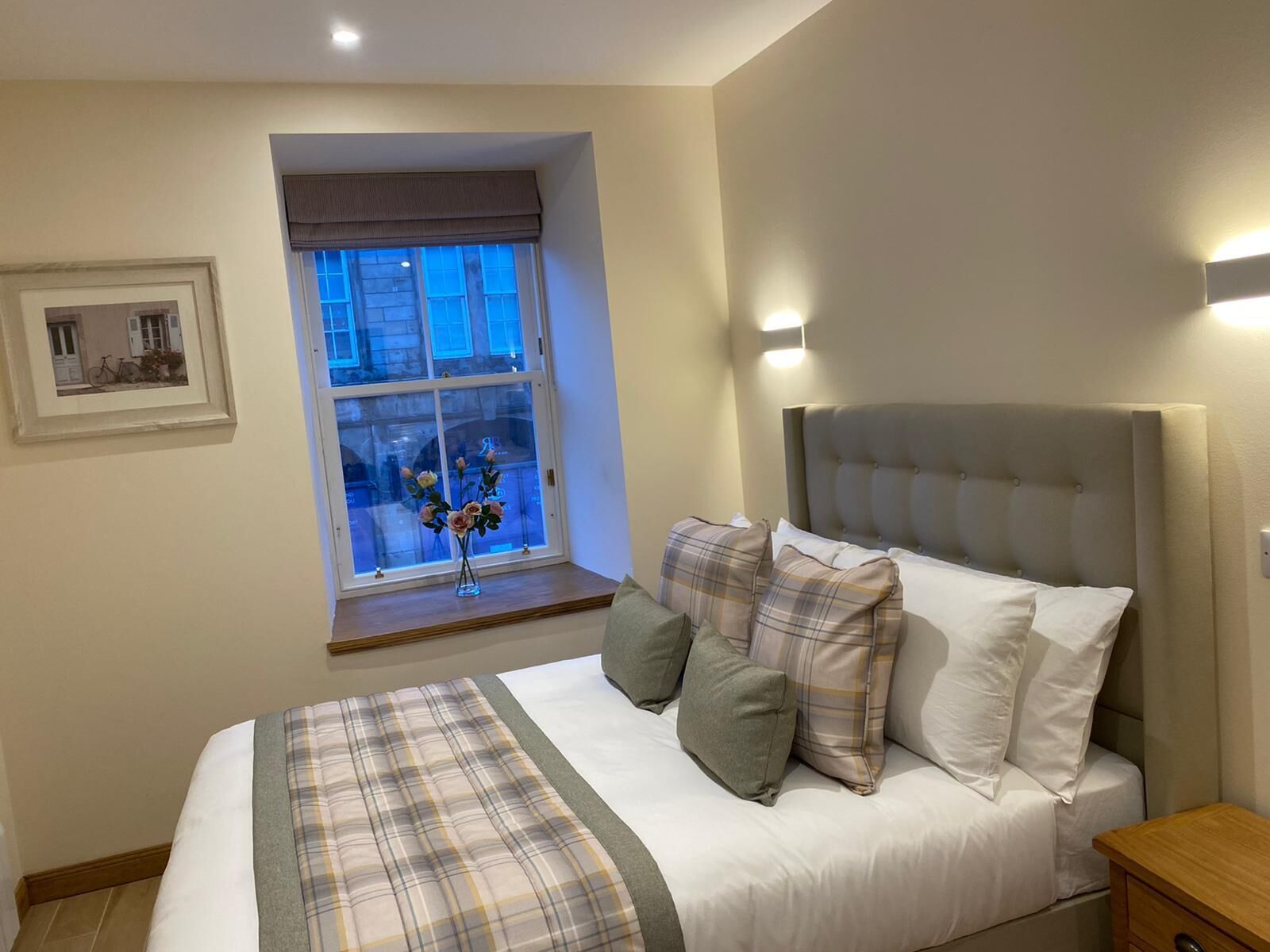 A bedroom with a bed, a window with a view, a bedside table, and a wall-mounted light.