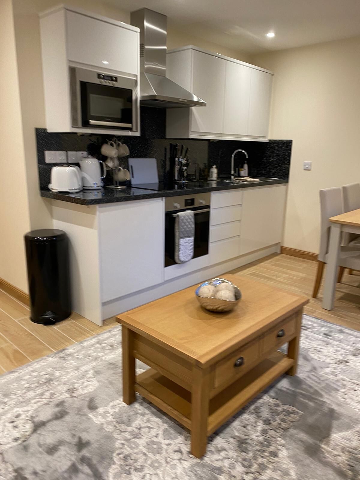 A compact kitchen with white cabinets, a black countertop, and a microwave. There's a small wooden table with a bowl on it.