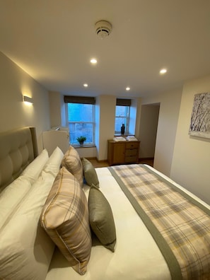 A bedroom with a large bed, two windows, a bedside table, and a wall-mounted artwork.