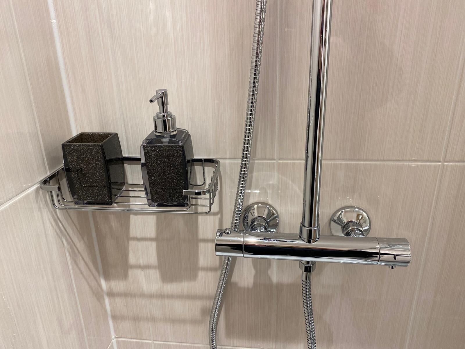 Una doccia con accessori cromati, un soffione a mano e due dispenser di sapone su uno scaffale.