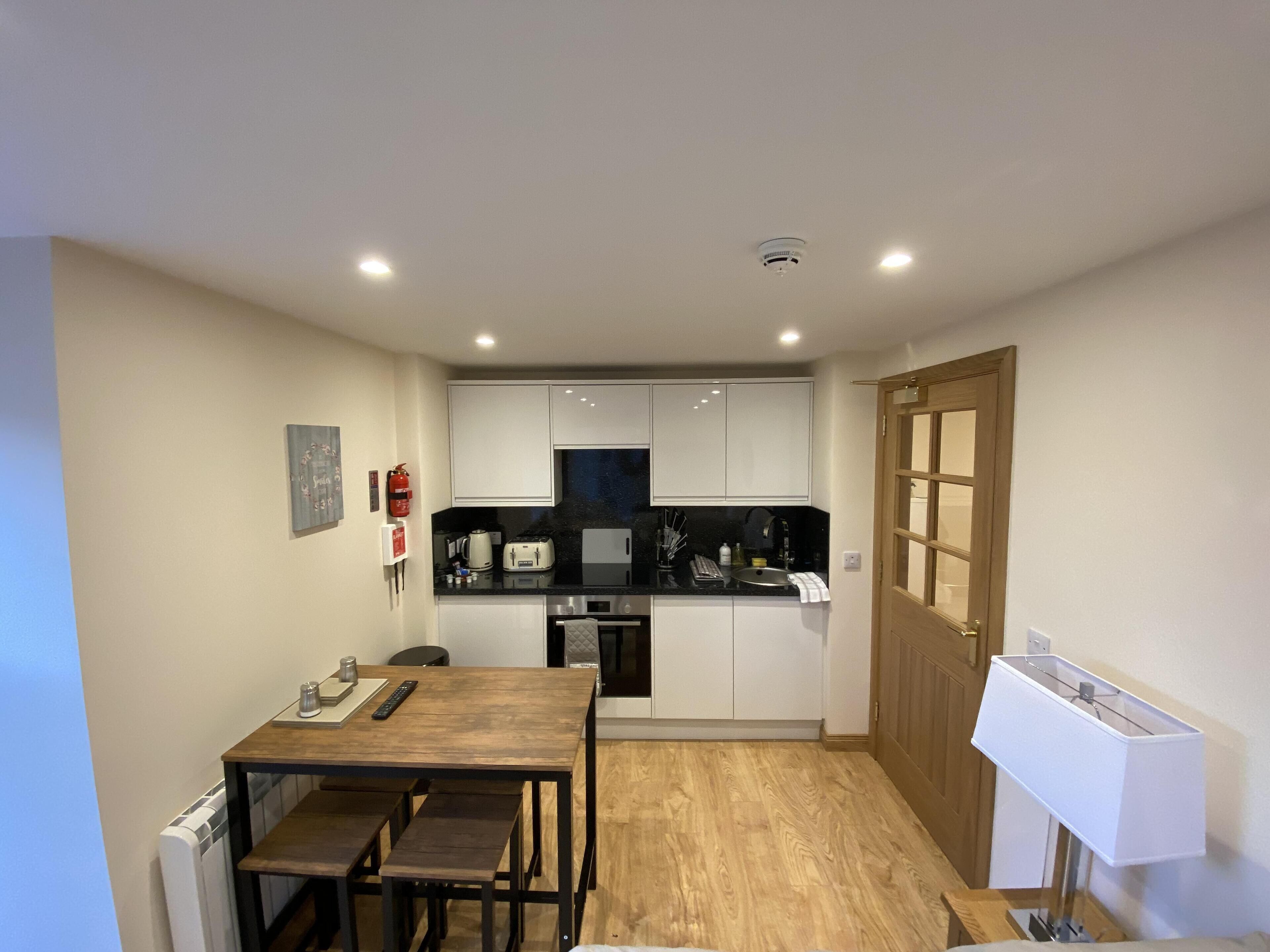 Une cuisine compacte avec des meubles blancs, une table à manger en bois avec des bancs, un four encastré et un luminaire suspendu blanc.