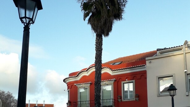 A street with a red building, a palm tree, and a lamp post. - Cozy Studio in the Historic Center of Cascais! (Cascais)