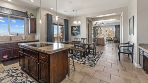 Eine großzügige Küche mit Granit-Arbeitsplatten, dunklen Holzschränken und einer großen Kochinsel. Es gibt einen Essbereich mit Tisch und Stühlen, sowie einen Blick ins Freie durch große Fenster.