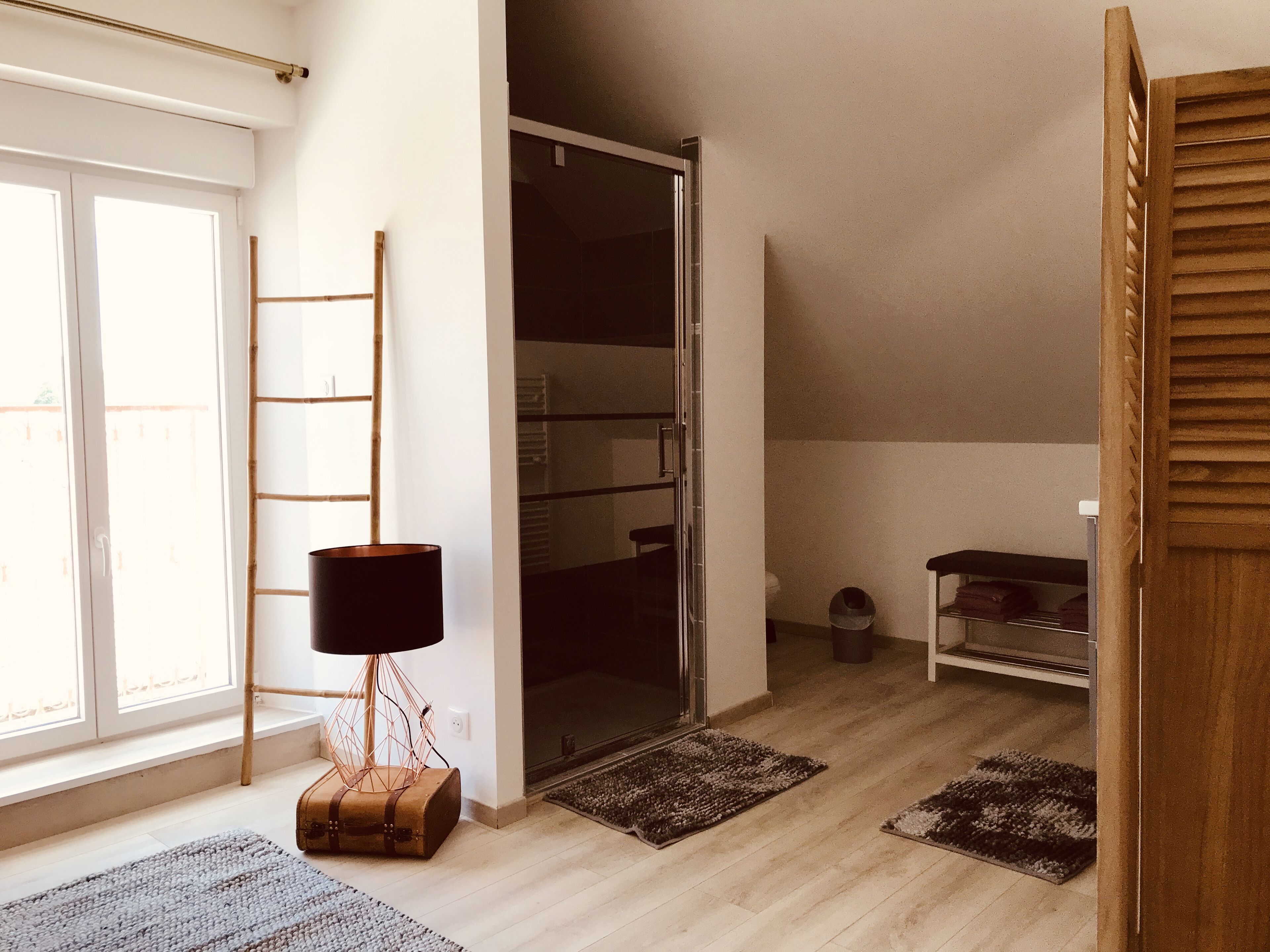 Una habitación con puerta de vidrio corrediza, escalera de madera, lámpara de pie, maleta y estante con ropa doblada.