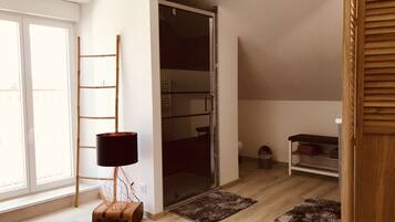 Una habitación con puerta de vidrio corrediza, escalera de madera, lámpara de pie, maleta y estante con ropa doblada.