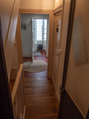 Un pasillo estrecho con suelo de madera, una puerta blanca a la derecha y un acceso al final que conduce a una habitación con ventana y escalera.