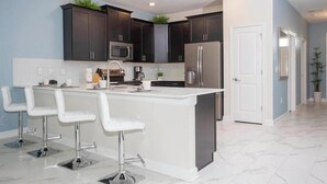 A modern kitchen with dark cabinets, a white island with bar stools, stainless steel appliances, and a tiled floor. - 1584mc-the Retreat at Championsgate (Davenport)