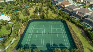 Luftaufnahme eines Tennisplatzes, umgeben von gepflegtem Rasen und Bäumen, mit Wohnhäusern im Hintergrund.