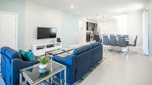 Un salon moderne avec un canapé d’angle, une table basse en verre et un téléviseur à écran plat. On y trouve également un coin repas avec une table et des chaises, ainsi qu’une cuisine en arrière-plan.