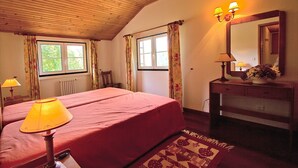 Un dormitorio con techo de madera, una cama con colcha roja, una mesita de noche con lámpara, un aparador con espejo y dos ventanas con cortinas.