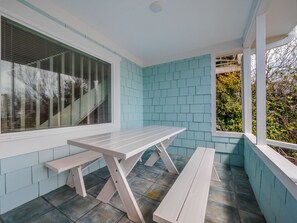 A covered patio area with a white picnic table and benches, light blue walls, and a view of greenery outside. - Surfside Cottage | Pet Friendly, Hot Tub, Views! (Topsail Beach)