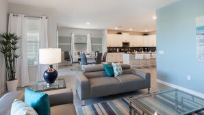 Ein modernes Wohnzimmer mit einem grauen Sofa, einem Glastisch und einem Streifenmuster-Teppich. Es gibt einen Essbereich mit Tisch und Stühlen sowie eine Küche mit weißen Schränken und einer Mikrowelle.