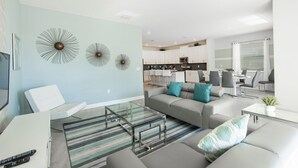 Een moderne woonkamer met een bank, een glazen salontafel en een gestreept vloerkleed. Achtergrond is een keuken met witte kasten en een eethoek met stoelen.