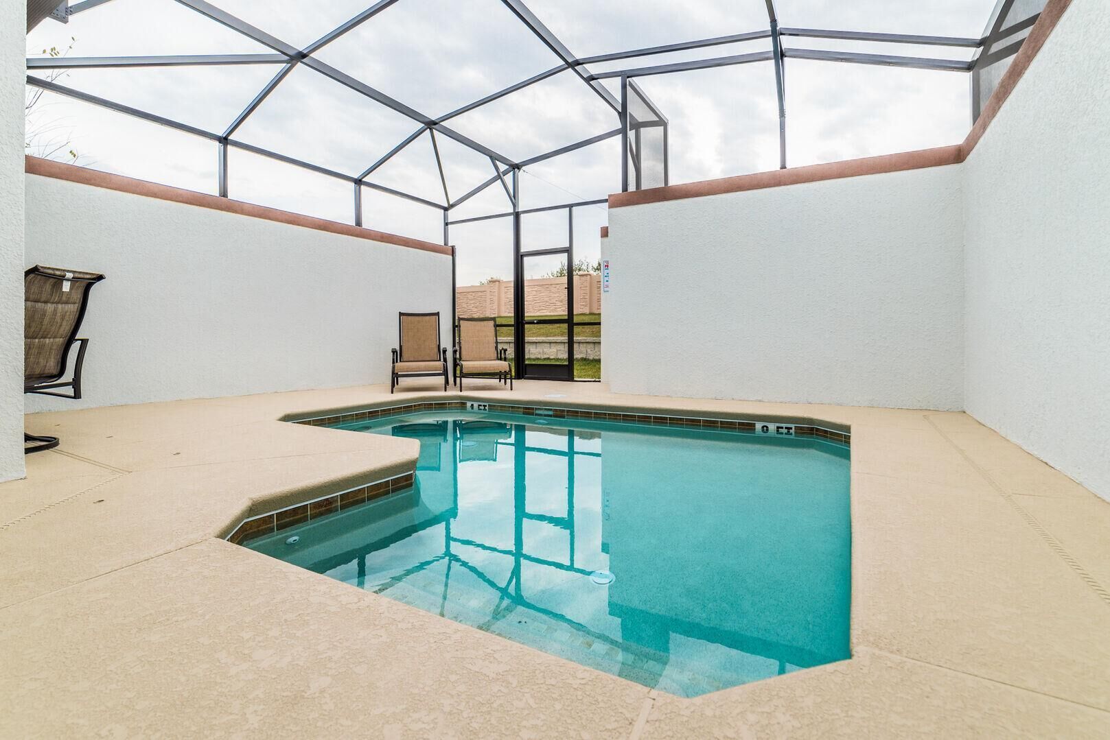 An indoor swimming pool with a glass ceiling, beige tiled floor, and two chairs near the pool.