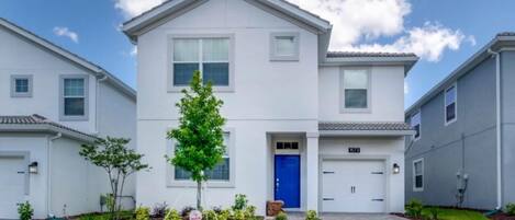 A modern two-story residential building with a blue front door, a small tree in the front yard, and a paved driveway.