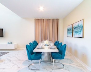 Een eethoek met een rechthoekige tafel voor zes personen, omringd door turquoise stoelen, een witte console en een beige gordijn.