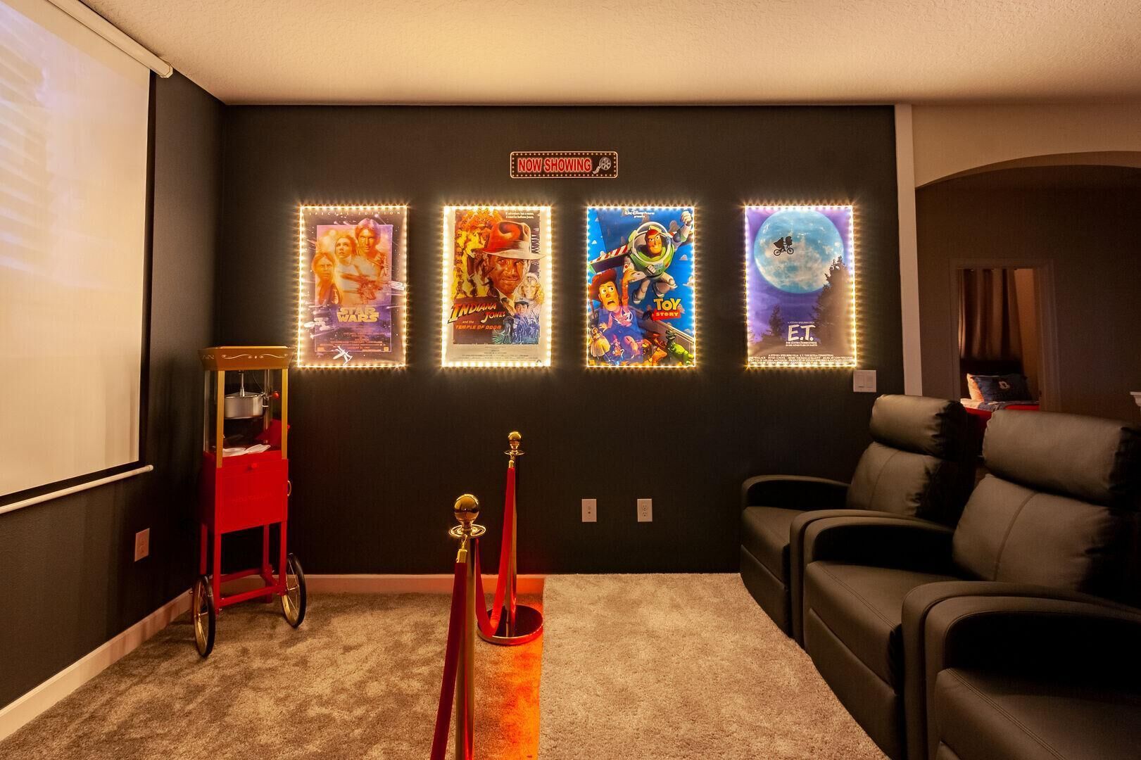 Una sala de cine en casa con una pared oscura, una pantalla de proyector y cuatro carteles de películas iluminados.