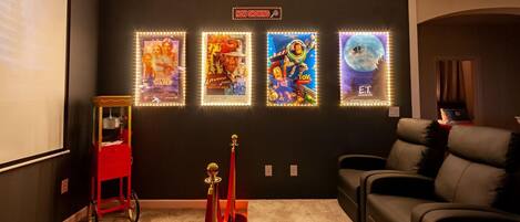 Una sala de cine en casa con una pared oscura, una pantalla de proyector y cuatro carteles de películas iluminados.