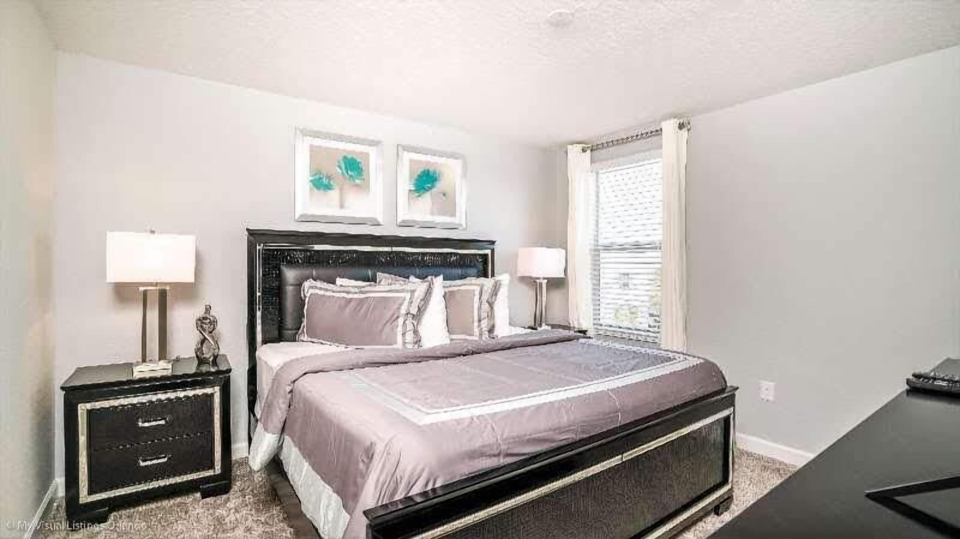 A bedroom with a bed, two bedside tables, a nightstand, a window with blinds, and two wall-mounted pictures.