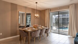 A dining area with a large table set for six, surrounded by six chairs with gold legs. There is a chandelier above the table and a large mirror on the wall. The room has a large sliding glass door leading to a pool area. - 840dsd-the Retreat at Championsgate (Kissimmee)
