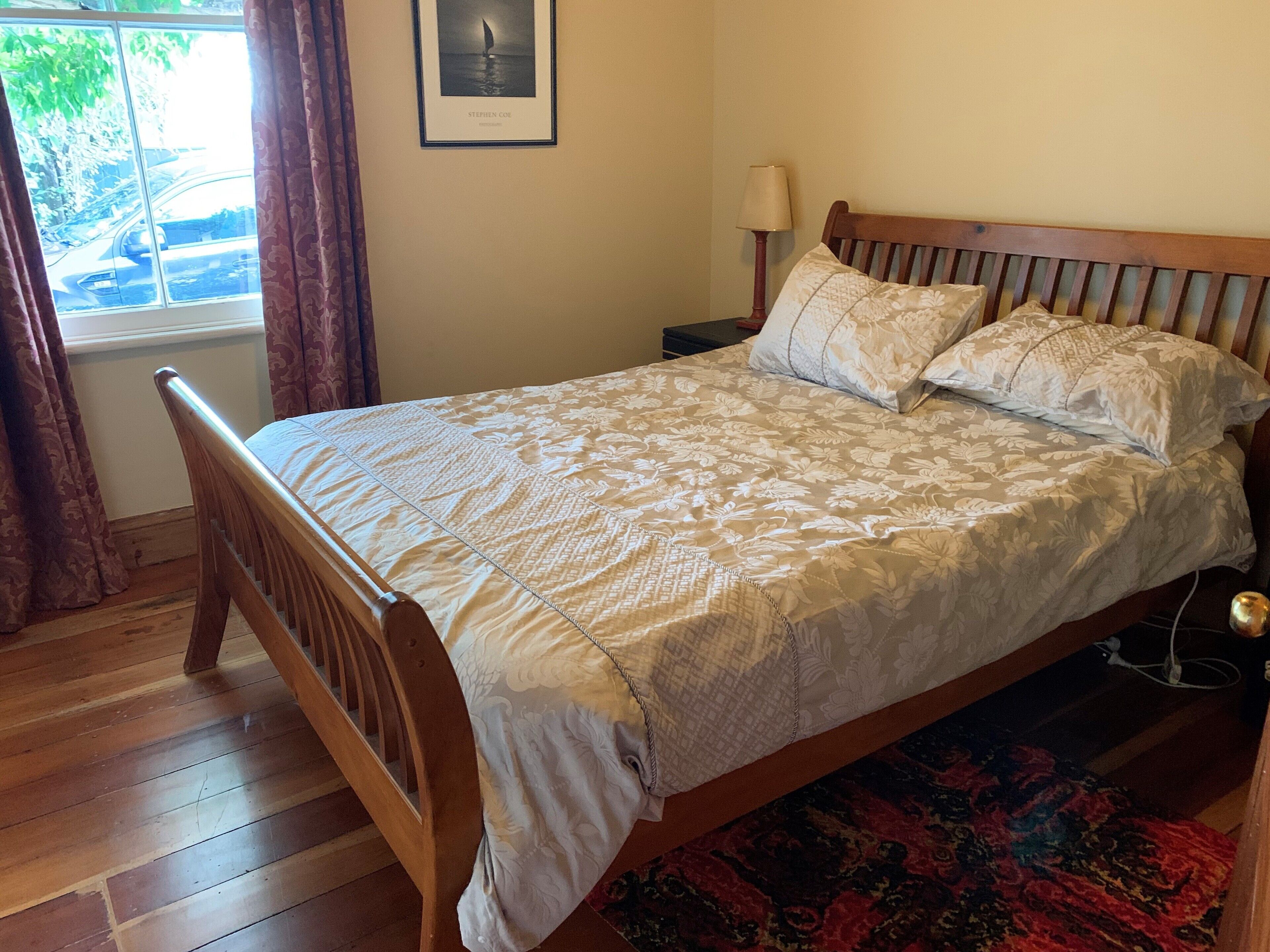 Un dormitorio con una cama de madera, un edredón estampado, una mesita de noche con una lámpara y una ventana con cortinas.
