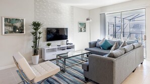 A modern living room with a grey sofa, a white entertainment unit, a flat-screen TV, and a large window offering a view of the outdoors. - 8912prw- NON Renting July 7, 2022 Windsor at Westside (Kissimmee)