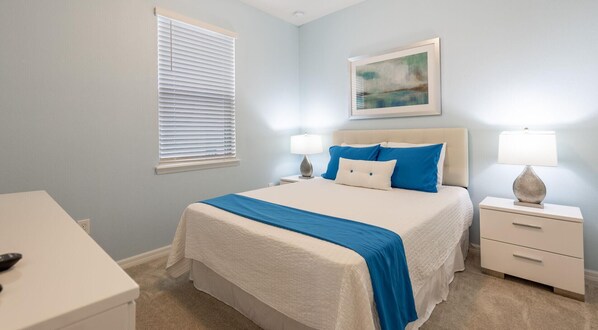 A bedroom with a bed, bedside tables, a nightstand, a lamp, a painting, and a window with blinds.