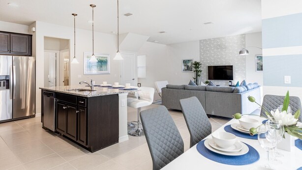 A modern kitchen with a central island, a dining area with blue placemats, and a living room with a gray sofa and a flat-screen TV. - 8912prw- NON Renting July 7, 2022 Windsor at Westside (Kissimmee)