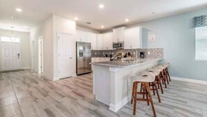 A modern kitchen with white cabinets, a large island with bar stools, stainless steel appliances, and a light blue accent wall. - C9036sid-the Retreat at Championsgate (Davenport)