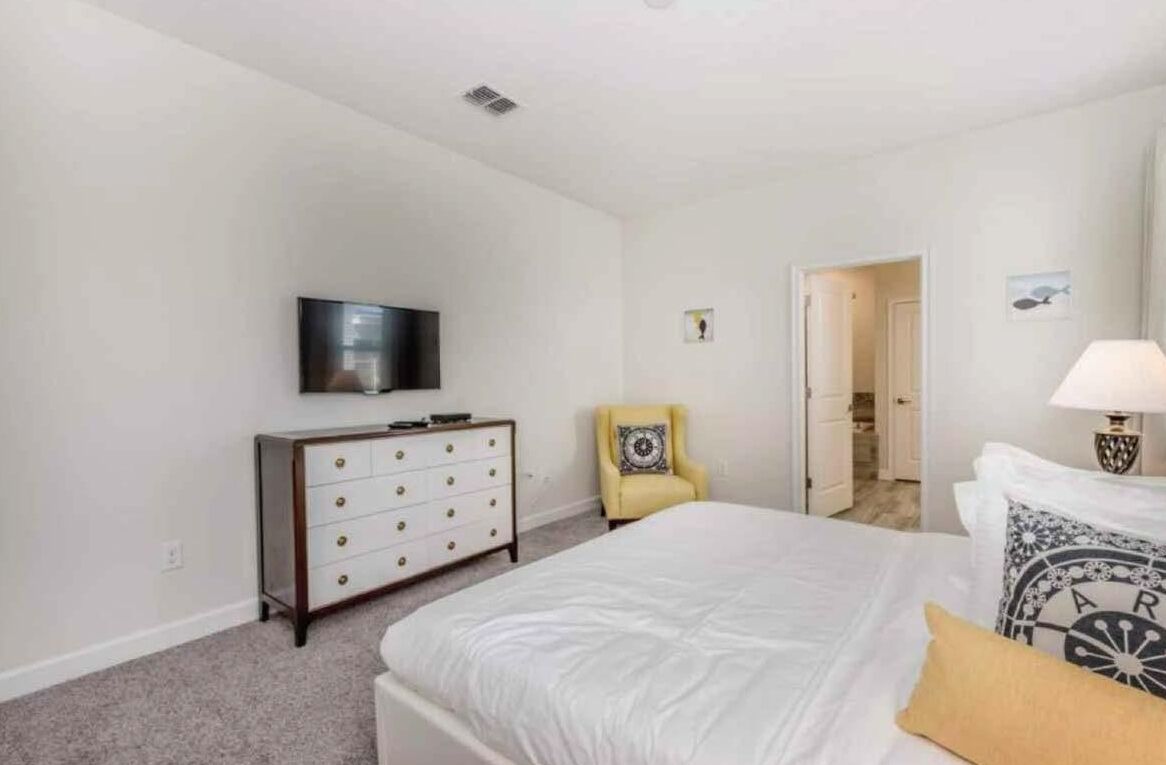 Une chambre à coucher comprenant un lit, une commode, un téléviseur, une chaise et une porte donnant sur une autre pièce.