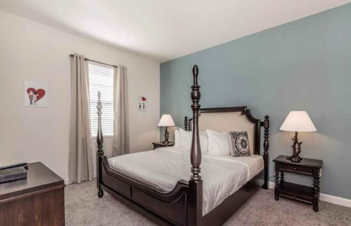 A bedroom with a four-poster bed, two bedside tables with lamps, a nightstand, a dresser, and a window with curtains.