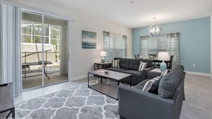 A modern living room with a gray sofa set, a coffee table, and a dining area visible through a glass sliding door. - C780pbd - The Retreat at Championsgate (Davenport)