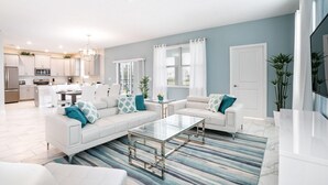 Una sala moderna con un sofá blanco, una mesa de centro de vidrio y un tapete a rayas. Hay una zona de cocina con gabinetes blancos y una mesa de comedor.