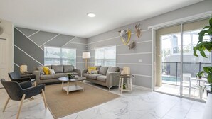A modern living room with two sofas, a coffee table, and a side table. There are large windows with blinds, a sliding glass door leading to a balcony, and a decorative wall piece.