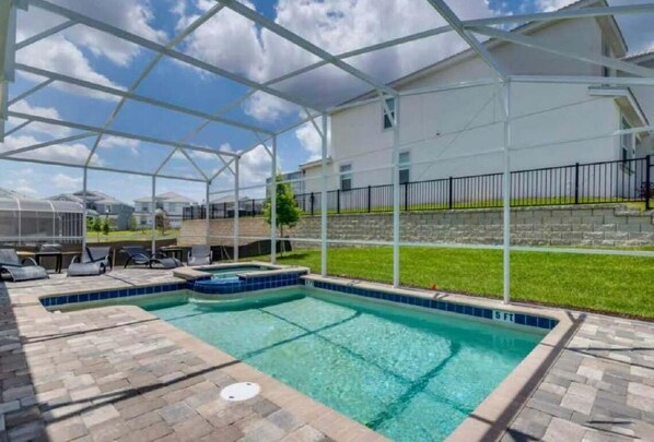 A covered swimming pool with a clear blue water feature, surrounded by a patio area with lounge chairs and a small hot tub.