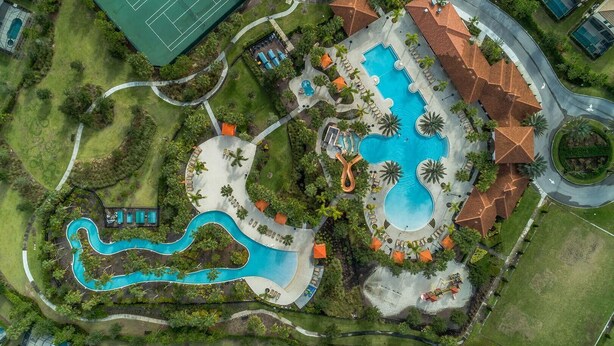 Vista aerea di un resort con una grande piscina centrale, un campo da tennis e una zona verde con palme.