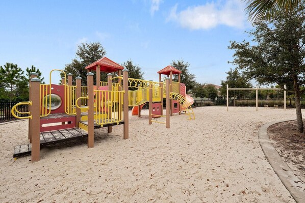 A playground with a sand surface, featuring a slide, climbing structures, and a small shelter.