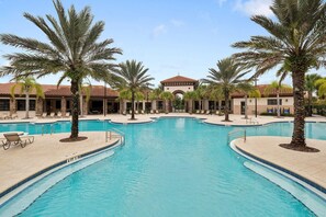 A large outdoor swimming pool surrounded by palm trees and lounge chairs, with a building in the background.
