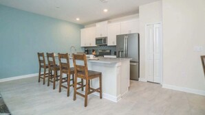 A modern kitchen with white cabinets, a stainless steel refrigerator, and a central island with bar stools. - C550pbd- The Retreat at Championsgate (Davenport)