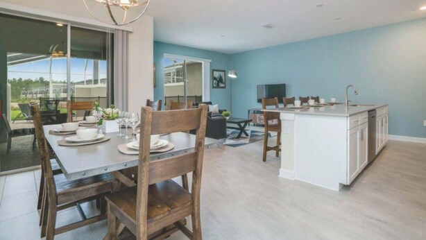 A modern kitchen with a central island, a dining area with a large table and chairs, and a view of the outdoors through a glass door. - C550pbd- The Retreat at Championsgate (Davenport)