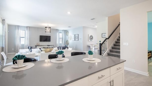 A modern kitchen with a central island, a dining area with white furniture, and a staircase leading to an upper floor. - 720dsd-the Retreat at Championsgate (Kissimmee)