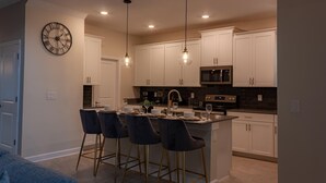 A modern kitchen with white cabinets, a central island with bar stools, pendant lighting, and a wall clock. - 801dsd-the Retreat at Championsgate (Kissimmee)
