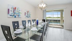 Un espace repas moderne avec une table en verre, des chaises noires et de l’assiette blanche. Les murs sont ornés de peintures abstraites et de grandes fenêtres offrent une vue sur une piscine.